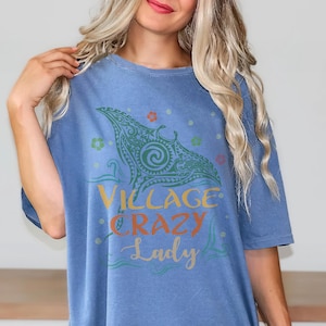 May include: A blue t-shirt with the words "Village Crazy Lady" in yellow, orange, and blue script. The shirt features a green tribal design with floral accents. The shirt is made of a soft material.