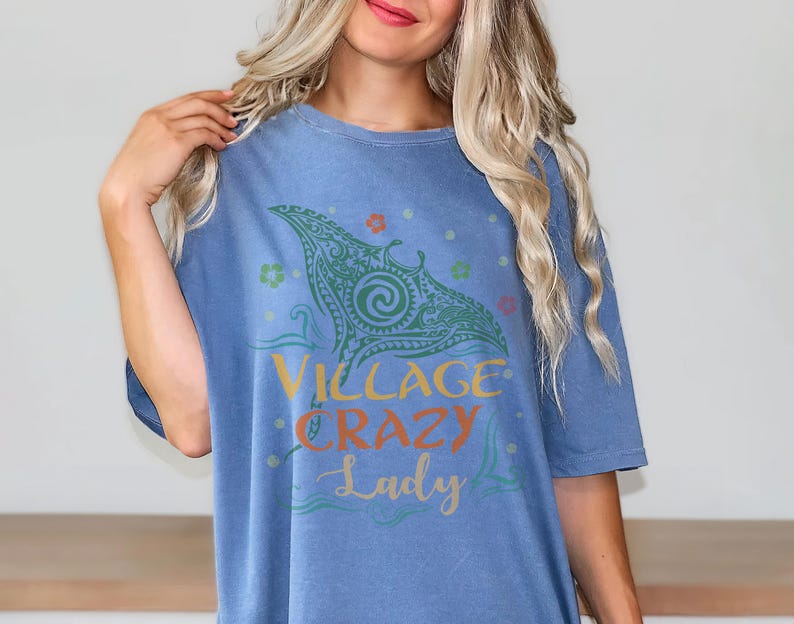 May include: A blue t-shirt with the words "Village Crazy Lady" in yellow, orange, and blue script. The shirt features a green tribal design with floral accents. The shirt is made of a soft material.