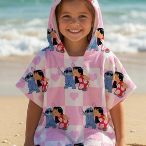 May include: A hooded poncho towel featuring a repeating pattern of Stitch and Lilo on a light pink and white checkered background with small pink hearts. The garment has short sleeves, a hood, and a soft texture, displaying the characters in a kiss.