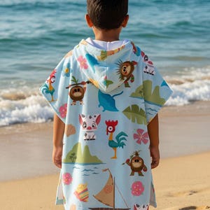 May include: A light blue hooded poncho towel featuring a repeating pattern of cartoon characters, including a rooster, a pig, and small coconut creatures, alongside tropical flowers, green islands, seashells, and a small wooden sailboat.