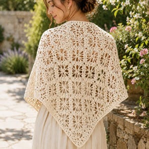 May include: A cream-colored triangular shawl featuring an intricate, repeating crochet pattern of interconnected floral motifs. The garment is draped over the shoulders, showcasing a delicate, openwork design with a pointed hem and scalloped edges.