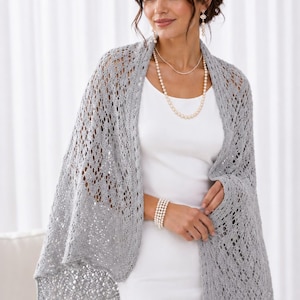 May include: A light grey, open-knit shawl featuring a delicate, repeating geometric lace pattern with a scalloped edge. The shawl is draped over a simple, sleeveless white dress, complemented by a multi-strand pearl necklace and a matching pearl bracelet.