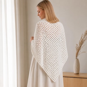 May include: A triangular off-white crochet shawl featuring a repeating openwork granny square pattern. The shawl is draped over the shoulders, showcasing a textured, lightweight yarn construction with a pointed hemline and scalloped edges.