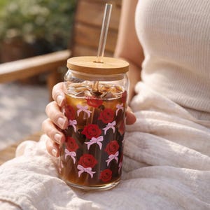 May include: A clear glass tumbler filled with iced tea, adorned with red roses and pink bows. It has a bamboo lid and a clear glass straw. The tumbler is held by a person, suggesting a refreshing beverage for outdoor enjoyment.