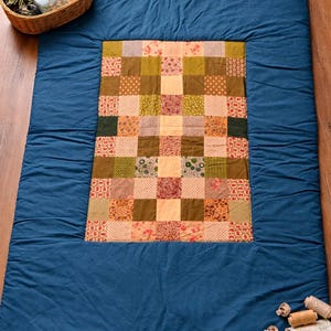 Puede incluir: Un edredón hecho a mano con un centro de patchwork en varias telas florales y estampadas, enmarcado por un borde azul liso. El edredón se exhibe sobre una superficie de madera, con una cesta de suministros de costura cerca.