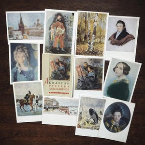 Op de afbeelding: Een verzameling vintage ansichtkaarten met aquarel schilderijen van Russische kunstenaars. De ansichtkaarten tonen portretten, landschappen en scènes uit de Russische geschiedenis. Een boek getiteld "Aquarellen van Russische kunstenaars" is ook aanwezig.