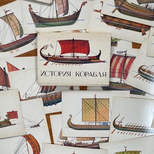 Puede incluir: Un conjunto de tarjetas vintage con ilustraciones de varios veleros con velas coloridas. Una caja con el texto cirílico "ИСТОРИЯ КОРАБЛЯ" está en el centro. Las tarjetas están dispuestas sobre una superficie de madera.