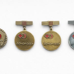 May include: A collection of vintage Soviet donor badges. The badges feature a red cross and crescent design, with the text "Donor USSR" in Cyrillic script. The badges are in gold and silver tones, with a variety of shapes and levels.