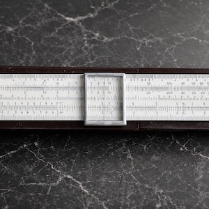 Könnte beinhalten: Ein Vintage-Rechenschieber mit weißen Skalen und einem braunen Rahmen. Der Rechenschieber hat einen transparenten Cursor und ist mit Zahlen und Maßen markiert. Es ist ein Werkzeug für mathematische Berechnungen.