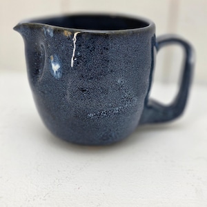 May include: A handmade, navy blue ceramic creamer with a handle and spout. The creamer has a speckled texture and a unique, organic shape. The interior is dark, and the exterior has a glossy finish. The creamer is approximately 4 inches tall.
