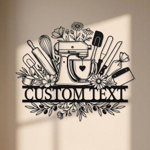 Op de afbeelding: Een zwart metalen wanddecoratie met een keukenmixer met een hart op de kom, omringd door bakgereedschap zoals een garde, deegroller en spatel, geaccentueerd met bloemmotieven. Onder de gereedschappen staat een banner met de tekst CUSTOM TEXT.