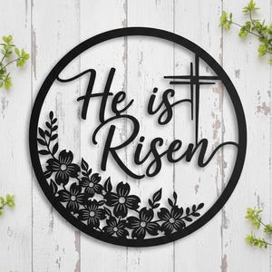 Könnte beinhalten: Schwarze Metall-Wandkunst mit dem Schriftzug "He is Risen" in eleganter Schrift, umrahmt von einem schwarzen Kreis. Ein Kreuz ist integriert, mit floralen Elementen am unteren Rand. Der Hintergrund ist weißes, verwittertes Holz.