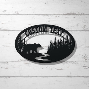 Könnte beinhalten: Schwarze Metall-Wandkunst mit einem Bären, der in einer Waldszene in der Nähe eines Flusses geht. Das Design wird von einer ovalen Form mit dem Text "CUSTOM TEXT" oben eingerahmt. Die Kunst ist auf einem weißen Holzhintergrund montiert.