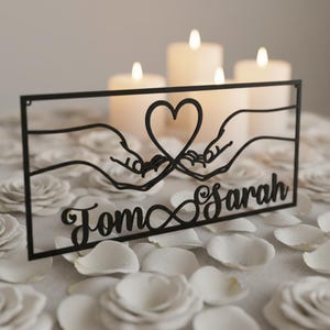 Könnte beinhalten: Schwarzes Metall-Wandkunstwerk mit einem Herzen, das von zwei Händen gebildet wird, mit den Namen "Tom & Sarah" darunter. Das Kunstwerk ist vor dem Hintergrund von brennenden Kerzen und weißen Rosenblättern platziert.