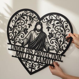 Könnte beinhalten: Schwarze Metall-Wandkunst in Herzform mit einer Silhouette von Jesus mit ausgestreckten Armen, umgeben von floralen und Kreuz-Designs. Der Text "UNDER HIS PROTECTION MILLER FAMILY" ist darunter zu sehen. Das Kunstwerk wird von zwei Händen gehalten.