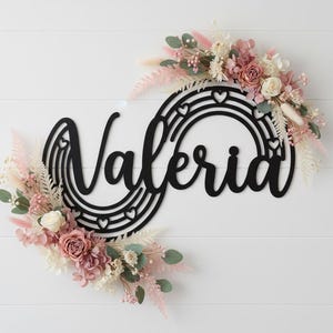 Könnte beinhalten: Ein schwarzes Holzschild mit dem Namen "Valeria" in Schreibschrift, eingebettet in ein Regenbogen-Design mit Herz-Ausschnitten. Das Schild ist mit einem Blumengesteck aus rosa, weißen und grünen Trockenblumen verziert und schafft eine dekorative Wanddekoration.