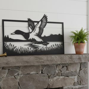 Könnte beinhalten: Schwarze Metall-Wandkunst, die eine Ente im Flug über einer See-Szene darstellt. Das Kunstwerk ist gerahmt und an einer weißen Wand über einem Steinkamin angebracht. Eine Topffarn und ein Messinghorn sind ebenfalls zu sehen.