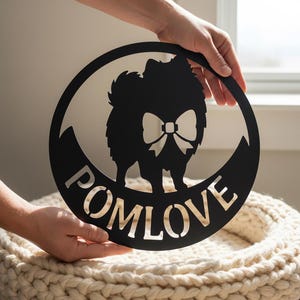 Könnte beinhalten: Schwarzes Metallschild mit dem Wort "POMLOVE" und der Silhouette eines Zwergspitzes mit Fliege. Das Schild ist rund und wird von Händen gehalten.