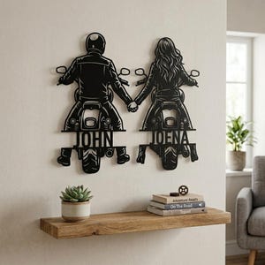 Op de afbeelding: Een zwart metalen wanddecoratie met twee figuren op motorfietsen die elkaars hand vasthouden. Het linkerfiguur is gelabeld met JOHN en het rechterfiguur met JOENA. Het ontwerp toont twee rijders van achteren, elk op hun eigen motor, met de namen geïntegreerd in het metalen frame onder de zadels.