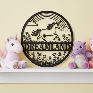 Könnte beinhalten: Ein schwarzes Metallschild mit der Aufschrift Dreamland, das ein Einhorn, einen Regenbogen und ein Blumendesign zeigt. Das runde Schild wird von rosa und lila Plüsch-Einhorn- und Drachenspielzeugen umgeben. Das Schild ist auf einem weißen Regal montiert.