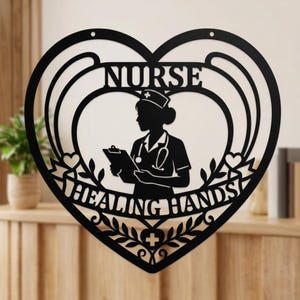 Könnte beinhalten: Schwarze Metall-Wandkunst in Herzform mit den Worten "NURSE" und "HEALING HANDS". Eine Silhouette einer Krankenschwester mit Klemmbrett und Stethoskop befindet sich in der Mitte. Das Design umfasst florale und Herz-Akzente.