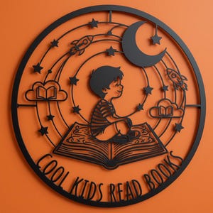 Könnte beinhalten: Schwarze Metall-Wandkunst mit einem Kind, das auf einem offenen Buch sitzt, umgeben von Sternen, Raketen und einem Halbmond. Der Text "COOL KIDS READ BOOKS" befindet sich unten. Das Kunstwerk ist kreisförmig.