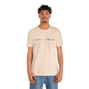 May include: A cream-coloured t-shirt with the text "coffee then... Wine" in a dark brown font. The shirt is a crew neck and is being worn by a person in blue jeans.