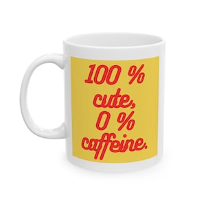 May include: White ceramic mug with a yellow rectangle featuring the text "100% cute, 0% caffeine" in red script. The mug has a curved handle and is designed for hot beverages.
