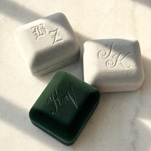 Peut inclure: Trois boîtes à bagues carrées en velours gris clair et vert foncé. Chaque boîte porte un monogramme en écriture script. Les boîtes sont disposées sur une surface claire, avec des ombres de la lumière du soleil.