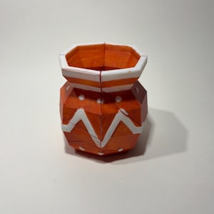 May include: A faceted orange vase with a white rim and white geometric patterns. The design features a series of white dots and a zigzag line encircling the body. The object has a distinct polygonal structure with visible seams between the panels.