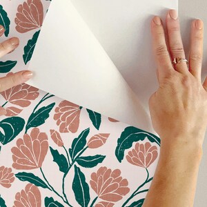 May include: Floral wallpaper with a repeating pattern of coral-colored flowers and dark green leaves on a white background. The wallpaper is being peeled back to reveal the wall behind it.