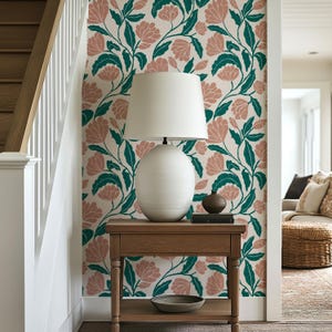 May include: A white ceramic lamp with a white lampshade sits on a wooden table. The table is in front of a wall with a floral pattern of pink flowers and green leaves on a cream background. A small decorative vase and a book are on the table.