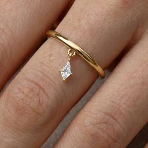 Könnte beinhalten: Ein goldener Ring mit einem kleinen, rautenförmigen Anhänger. Der Ring ist schlicht und elegant, mit einem dünnen Band und einem zarten Anhänger. Der Anhänger ist ein klarer, funkelnder Edelstein.
