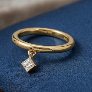 May include: A gold ring with a small, square, clear gemstone pendant. The ring is set against a blue surface, with a wooden and marble background. The ring is a simple band with a dangling charm.