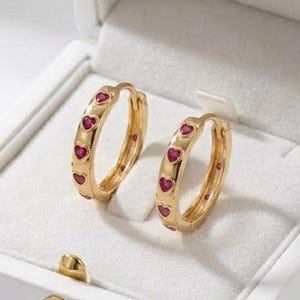 May include: A pair of gold-colored hoop earrings with small, heart-shaped, red gemstones. The earrings are displayed inside a white jewelry box with a gold-colored clasp.