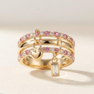 May include: A gold ring with three bands, each adorned with pink and white gemstones. Two charms dangle from the ring: a heart and a rectangular stone. The ring is set against a soft, neutral background.