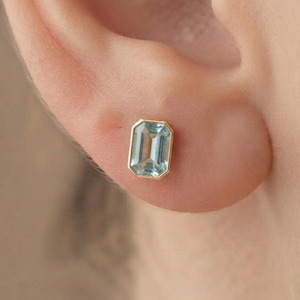 May include: A single stud earring featuring a rectangular emerald-cut light blue gemstone set in a polished gold bezel setting, securely positioned on the earlobe with a clean and simple design aesthetic.