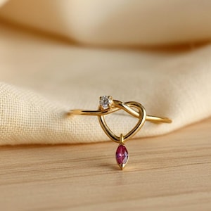May include: A gold-colored ring with a heart-shaped design. The ring features a small, clear gemstone at the top and a dangling, teardrop-shaped, red gemstone. The ring is set against a neutral background.