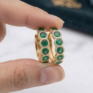 May include: A pair of gold hoop earrings featuring a row of round, vibrant green gemstones set in individual bezels. Each green stone is surrounded by a delicate halo of tiny, clear pavé-set stones, creating a textured, circular design.