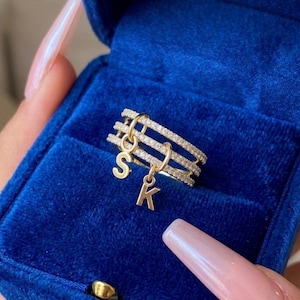 Pode incluir: Um anel dourado com três faixas, cada uma incrustada com pequenas pedras transparentes. Dois pingentes dourados, um com a letra "S" e outro com "K", pendem do anel. O anel é apresentado em uma caixa de joias de veludo azul.