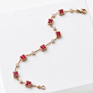 May include: A gold-toned chain bracelet featuring alternating square-cut red gemstones and small round clear stones. The bracelet is constructed with a delicate metal link chain and includes a secure lobster claw clasp at one end for closure.