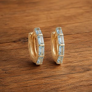 Puede incluir: Un par de pendientes de aro dorados con una hilera de piedras preciosas de color azul claro en talla baguette engastadas a lo largo de la curva frontal de cada aro. Los pendientes tienen un mecanismo de cierre de bisagra y acabado pulido.