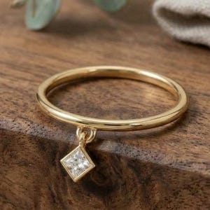 Pode incluir: Um anel fino de ouro com um pequeno pingente de diamante em forma de losango pendurado na banda. O metal tem um acabamento polido e brilhante, e a pedra transparente e facetada está engastada numa moldura de ouro simples.