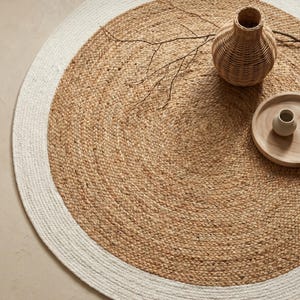 May include: A round jute rug with a white border. The rug has a natural, woven texture and is adorned with a woven vase, a small wooden tray, and a decorative branch. The rug's diameter is approximately 60 inches.