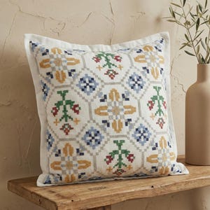 Könnte beinhalten: Quadratisches weißes Kissen mit einem farbenfrohen geometrischen Muster. Das Muster zeigt blaue, gelbe, grüne und rote Kreuzstichmuster auf weißem Hintergrund. Das Kissen hat einen weißen Rand und liegt auf einer Holzbank. Misst ca. 40 cm x 40 cm.