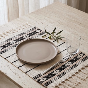 Op de afbeelding: Een rechthoekige geweven placemat met een crèmekleurige basis, zwarte en beige geometrische patronen en franjes. Op de placemat staan een taupekleurig keramieken bord, een helder glazen beker, een zilveren metalen vork en een kleine olijftak.