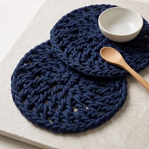 Puede incluir: Dos posavasos circulares de color azul marino hechos de cuerda gruesa trenzada con un patrón calado. Un pequeño cuenco de cerámica blanco roto moteado y una cuchara de madera clara están colocados sobre los posavasos de diseño tejido.