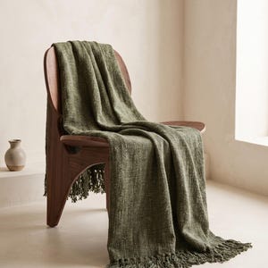 May include: A textured olive green throw blanket with fringe detailing draped over a curved wooden chair. The fabric features a woven appearance with subtle variations in color throughout the material, creating a consistent look across the surface.