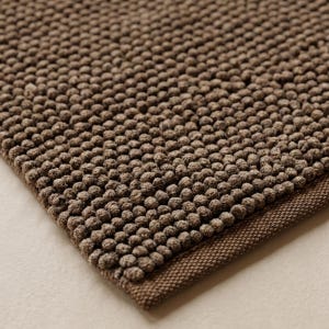 May include: A brown bath mat featuring a dense, textured surface composed of numerous small, rounded, pebble-like tufts. The mat has a flat, woven border along the edge, providing a clean finish to the overall design of the rectangular floor covering.