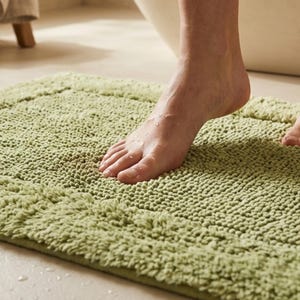 Può includere: Un tappetino da bagno rettangolare verde chiaro con una superficie strutturata e ad anello. Il tappetino è posizionato su un pavimento chiaro, con un piede che ci cammina sopra. Goccioline d'acqua sono visibili sul piede e sul tappetino.